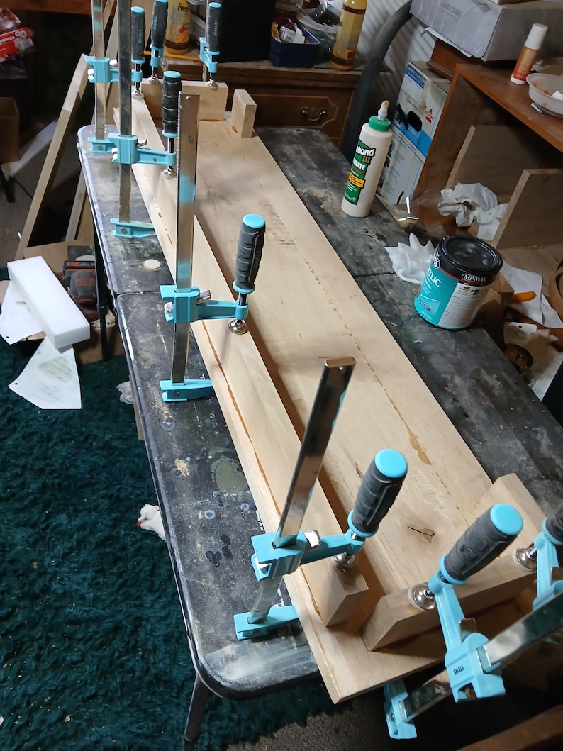 May include: A woodworking project with a long wooden plank held together by several blue and black clamps. The clamps are securing the wood on a dark surface. Tools and supplies are visible in the background.