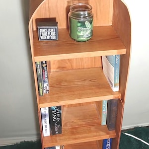 May include: A wooden bookshelf with a curved top and four shelves. The top shelf holds a candle in a glass jar and a small black box with the text "SORRY, I CAN'T I HAVE PLANS WITH MY DOG". The shelves contain books.