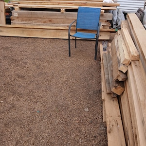 May include: A blue outdoor chair with a woven seat and back, set against a backdrop of stacked wooden planks and beams. The chair has a dark metal frame. The ground is covered in small brown pebbles.