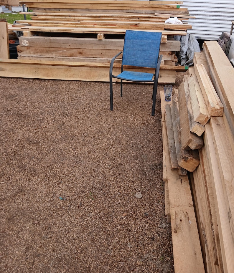 May include: A blue outdoor chair with a woven seat and back, set against a backdrop of stacked wooden planks and beams. The chair has a dark metal frame. The ground is covered in small brown pebbles.