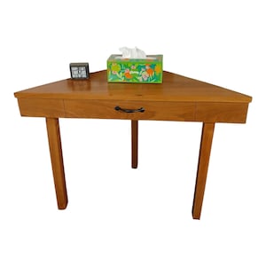 May include: A wooden corner desk with three legs and a drawer. A box of tissues and a small black sign that reads "SORRY I CAN'T HAVE PLANS WITH MY DOG" are on the desk.