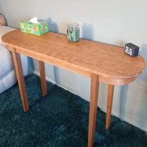 Puede incluir: Una consola de madera marr&oacute;n claro con una parte superior ovalada y cuatro patas delgadas. Una caja de pa&ntilde;uelos, un tarro con una vela y un peque&ntilde;o bloque negro con texto est&aacute;n sobre la mesa. La mesa est&aacute; sobre una alfombra verde azulado. La mesa mide unos 120 cm de largo.