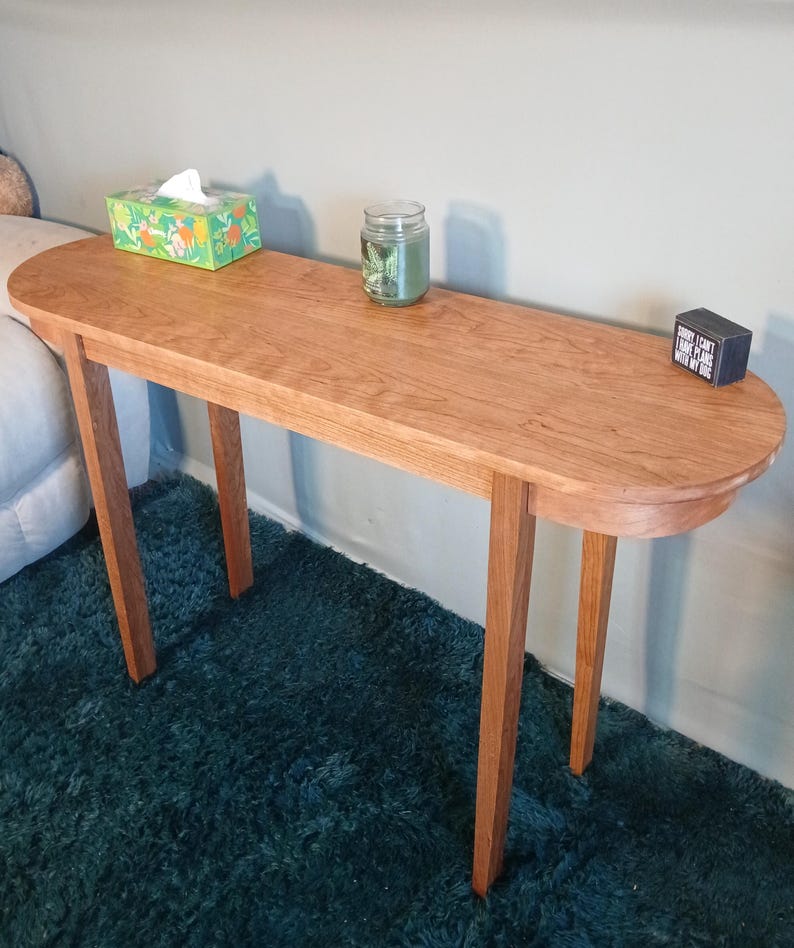 Puede incluir: Una consola de madera marr&oacute;n claro con una parte superior ovalada y cuatro patas delgadas. Una caja de pa&ntilde;uelos, un tarro con una vela y un peque&ntilde;o bloque negro con texto est&aacute;n sobre la mesa. La mesa est&aacute; sobre una alfombra verde azulado. La mesa mide unos 120 cm de largo.