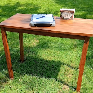 May include: A rectangular wooden table with a medium brown finish and four straight legs. On the surface sits a gray clipboard with white paper and a blue pen, alongside a small rectangular sign featuring the text CIRCA 1881 in black font.
