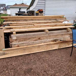May include: A pile of untreated wooden planks of various sizes, possibly for building or carpentry. The light-coloured wood shows visible grain. The planks are stacked outside, indicating a supply of timber, possibly in centimetres.
