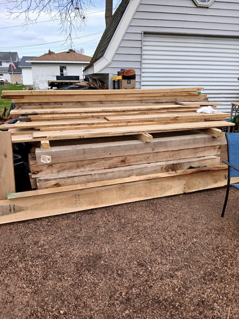 May include: A pile of untreated wooden planks of various sizes, possibly for building or carpentry. The light-coloured wood shows visible grain. The planks are stacked outside, indicating a supply of timber, possibly in centimetres.