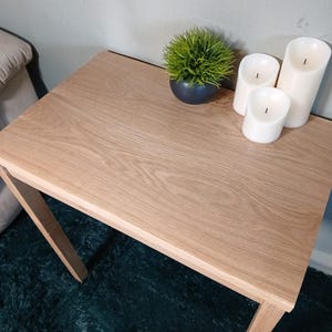 May include: A light oak side table with a square top and angled legs. The table is decorated with a small, round, dark blue pot with a green plant and three white pillar candles. The table is placed on a dark green rug.