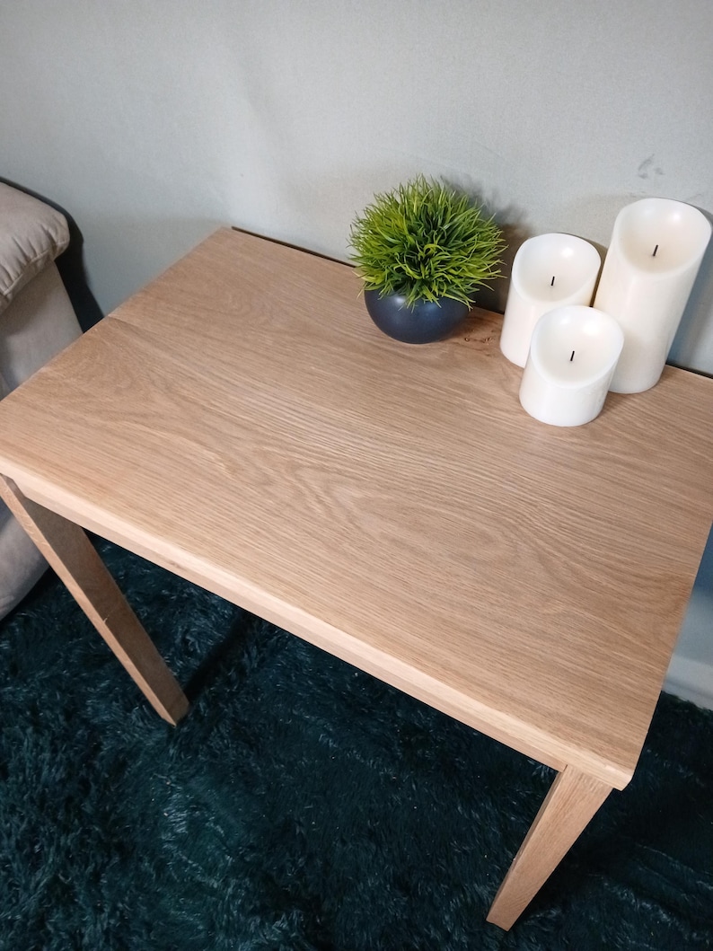 May include: A light oak side table with a square top and angled legs. The table is decorated with a small, round, dark blue pot with a green plant and three white pillar candles. The table is placed on a dark green rug.