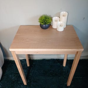 May include: A small, light-coloured wooden side table with a rectangular top and angled legs. The table is adorned with a small, dark blue pot containing green grass and three white pillar candles. The table is approximately 61 cm long.