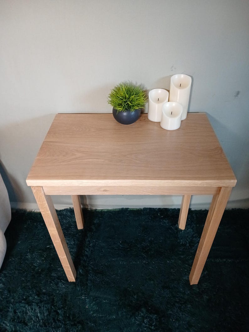 May include: A small, light-coloured wooden side table with a rectangular top and angled legs. The table is adorned with a small, dark blue pot containing green grass and three white pillar candles. The table is approximately 61 cm long.