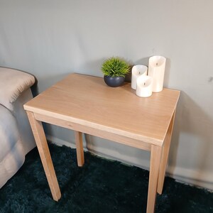 May include: A small, light-coloured wooden side table with a square top and four angled legs. The table has a small potted plant and three white pillar candles. The table is approximately 61 cm tall.