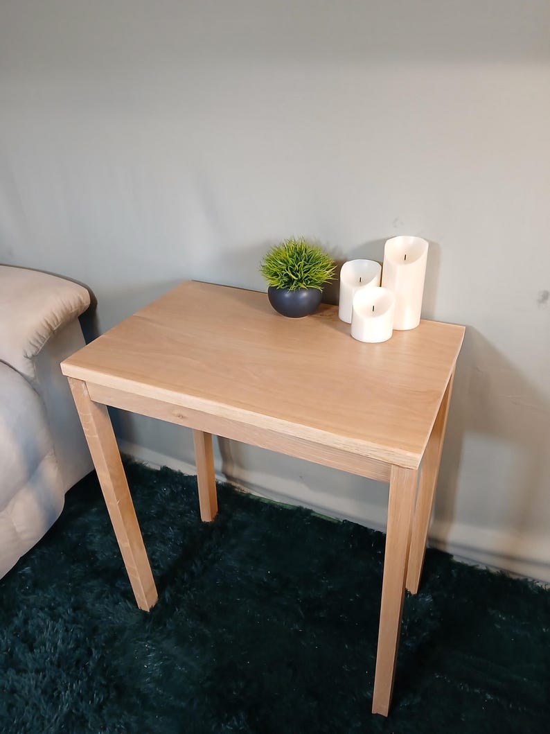 May include: A small, light-coloured wooden side table with a square top and four angled legs. The table has a small potted plant and three white pillar candles. The table is approximately 61 cm tall.