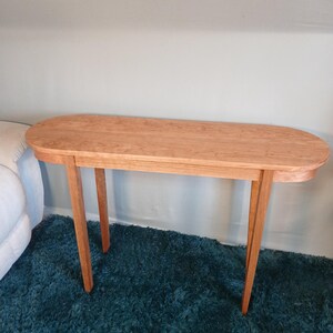 Puede incluir: Una consola de madera marr&oacute;n claro con una parte superior ovalada y cuatro patas anguladas. La mesa est&aacute; hecha de madera maciza y tiene un acabado natural. La mesa est&aacute; colocada sobre una alfombra de pelo largo color turquesa.