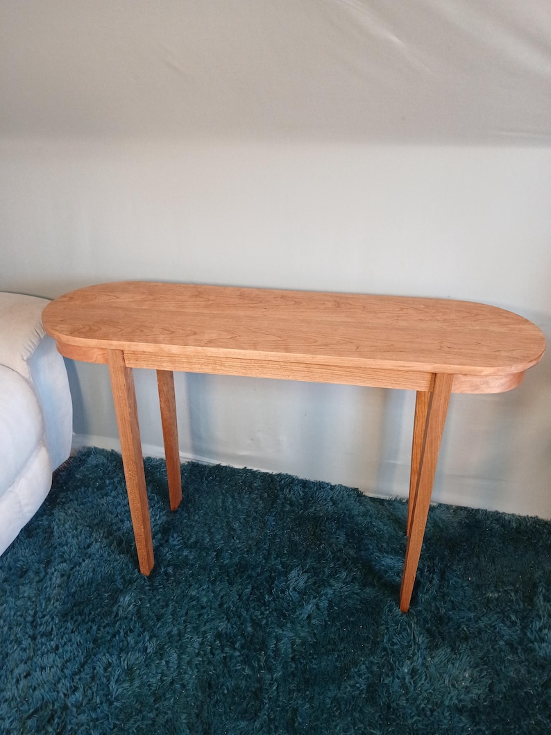 Puede incluir: Una consola de madera marr&oacute;n claro con una parte superior ovalada y cuatro patas anguladas. La mesa est&aacute; hecha de madera maciza y tiene un acabado natural. La mesa est&aacute; colocada sobre una alfombra de pelo largo color turquesa.