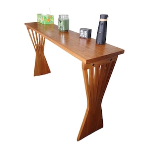 May include: A wooden console table with a unique design. The table has a long, rectangular top and two decorative legs. The legs are made of vertical wooden slats that fan out at the base. The table is made of a light brown wood and has a smooth finish.