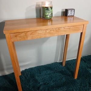 May include: A small, rectangular wooden table with a warm, natural finish. A lit candle in a glass jar and a small black box with white text are on the tabletop. The table is supported by four angled legs.