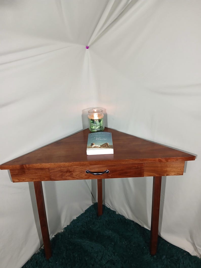 May include: A triangular wooden side table with a drawer and black handle. A book and a lit candle in a glass jar sit on top. The table has three legs and is made of dark wood.