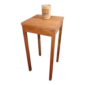 May include: A small, square wooden side table with four slender legs. A glass jar candle with a label that reads "Rich Coffee Warm Glow" sits on top. The table is made of light brown wood.