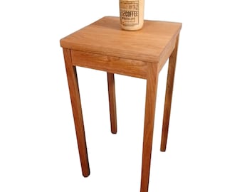 May include: A small, square wooden side table with four slender legs. A glass jar candle with a label that reads "Rich Coffee Warm Glow" sits on top. The table is made of light brown wood.