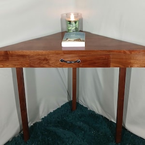 May include: A triangular wooden corner table with a drawer and black handle. The table has three legs and a rich, warm brown finish. A book and a lit candle in a glass jar sit on top. The table is approximately 30 inches wide.