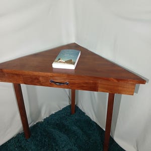 May include: A triangular wooden table with a drawer and three legs. A book with a woman on the cover is on the table. The table is a rich brown color and sits on a teal rug.