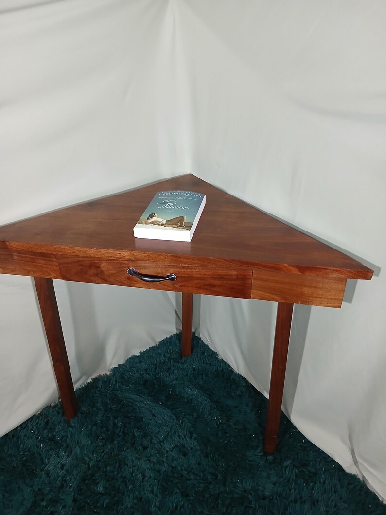 May include: A triangular wooden table with a drawer and three legs. A book with a woman on the cover is on the table. The table is a rich brown color and sits on a teal rug.