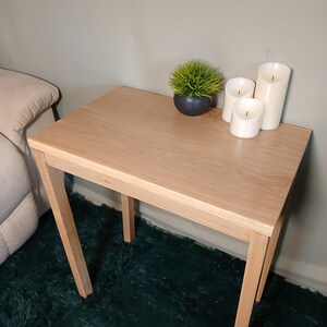 May include: A small, light-coloured wooden side table with a square top and four angled legs. The table is decorated with a small potted plant and three white pillar candles. The table is approximately 61 cm tall.