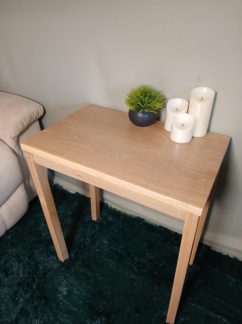 May include: A small, light-coloured wooden side table with a square top and four angled legs. The table is decorated with a small potted plant and three white pillar candles. The table is approximately 61 cm tall.