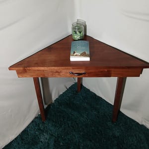 May include: A triangular, dark wood corner table with a drawer and three legs. A book titled "Maine" and a small jar are on the table. The table is set on a teal rug. The table is approximately 30 inches wide.