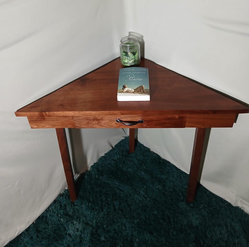 May include: A triangular, dark wood corner table with a drawer and three legs. A book titled "Maine" and a small jar are on the table. The table is set on a teal rug. The table is approximately 30 inches wide.