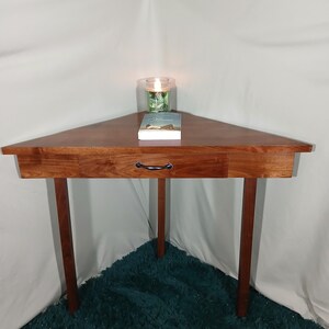 May include: A triangular wooden side table with a drawer and three legs. A book and a candle in a glass jar sit on top. The table is made of dark wood and is placed in a corner.