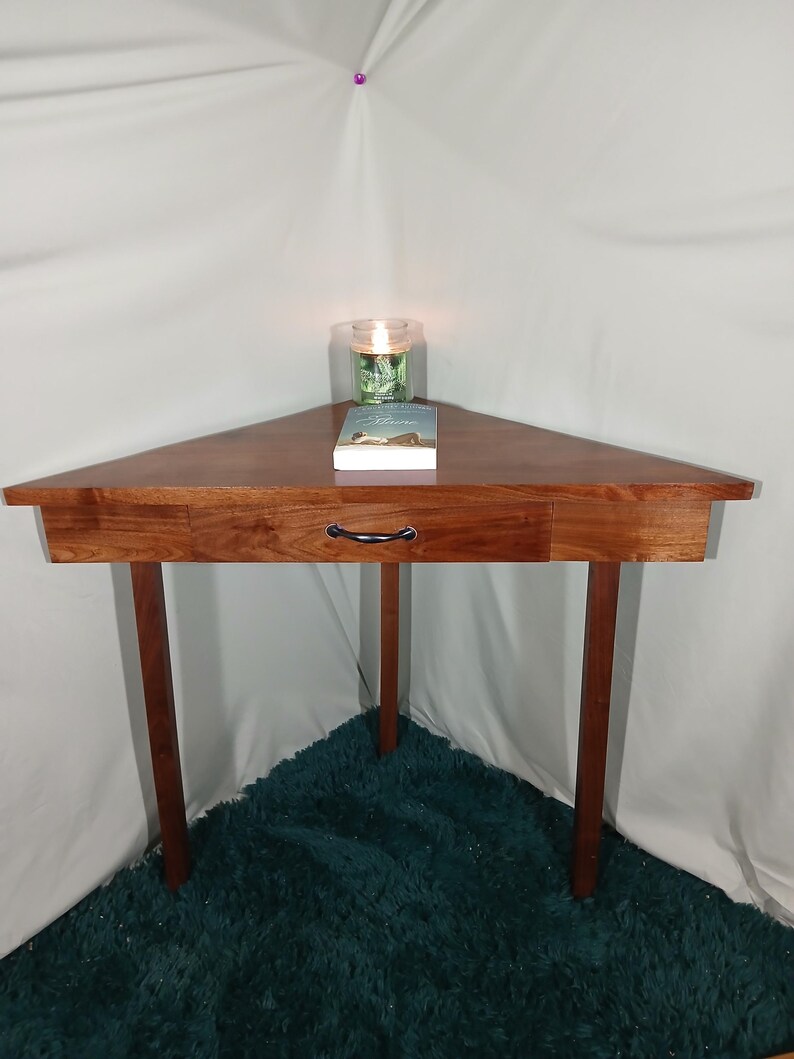 May include: A triangular wooden side table with a drawer and three legs. A book and a candle in a glass jar sit on top. The table is made of dark wood and is placed in a corner.