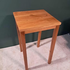 Peut inclure: Une petite table carrée en bois avec quatre pieds. La table est en bois brun clair et a une finition lisse et polie.