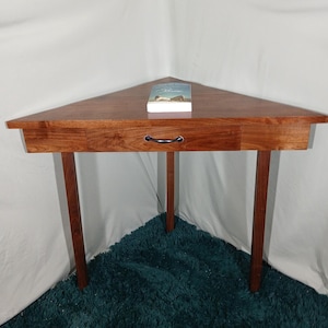 May include: A triangular wooden corner table with a drawer and three legs. The table is made of a reddish-brown wood and has a black handle on the drawer. A book rests on the table's surface. The table is approximately 30 inches wide.