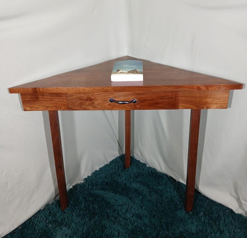 May include: A triangular wooden corner table with a drawer and three legs. The table is made of a reddish-brown wood and has a black handle on the drawer. A book rests on the table's surface. The table is approximately 30 inches wide.