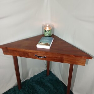 May include: A triangular wooden side table with a drawer and four legs. A book and a lit candle in a glass jar sit on the table's surface. The table is made of dark wood and is set on a teal rug.