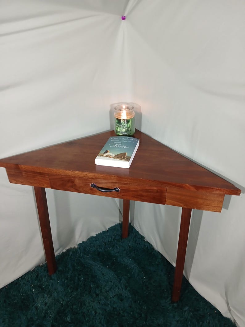 May include: A triangular wooden side table with a drawer and four legs. A book and a lit candle in a glass jar sit on the table's surface. The table is made of dark wood and is set on a teal rug.