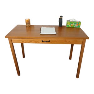 May include: A wooden desk with a drawer and angled legs. A container of coffee, a notebook, a black object, and a tissue box are on the desk. The desk is made of light brown wood.