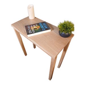 May include: A light oak-colored side table with a triangular top and angled legs. A book titled "The Bad Weather Friend" by Dean Koontz, a white candle, and a small potted plant are on the table.