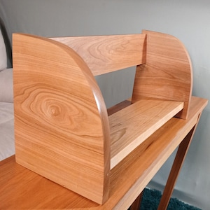 May include: A light brown wooden shelf with curved sides and a rectangular shelf. The shelf is made of light-colored wood and has a natural wood grain pattern. The shelf is sitting on a matching wooden table.