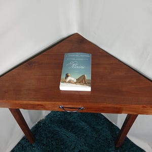 May include: A wooden, triangular-shaped table with a book titled "Maine" on top. The book cover features a woman on a beach. The table has a dark brown finish and a metal handle. The table is supported by angled legs.