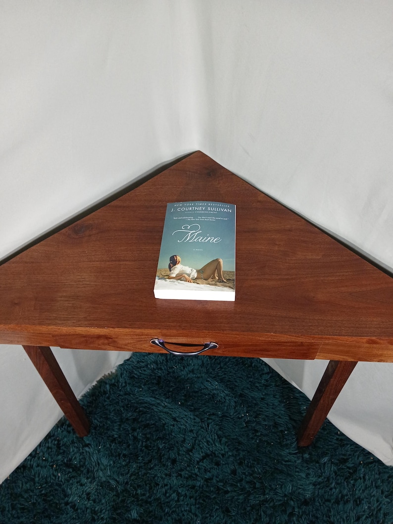 May include: A wooden, triangular-shaped table with a book titled "Maine" on top. The book cover features a woman on a beach. The table has a dark brown finish and a metal handle. The table is supported by angled legs.