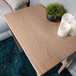 May include: A light oak square side table with a natural wood grain finish. The table is 61 cm wide and 61 cm long. A small black pot with green grass and three white pillar candles are on the table.