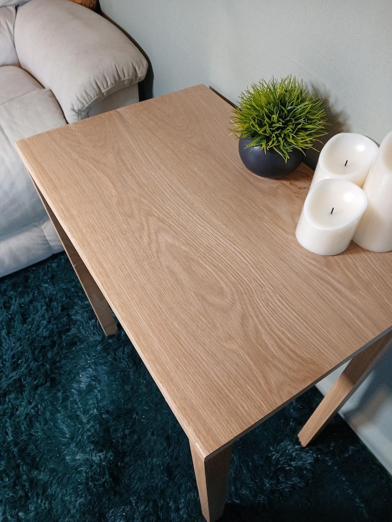 May include: A light oak square side table with a natural wood grain finish. The table is 61 cm wide and 61 cm long. A small black pot with green grass and three white pillar candles are on the table.