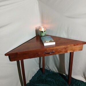 May include: A triangular wooden side table with a drawer and black handle. The table is a rich brown color and has four slender legs. A lit candle and a book are on top of the table.