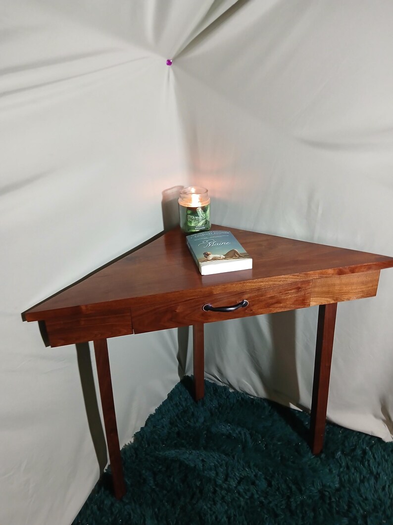 May include: A triangular wooden side table with a drawer and black handle. The table is a rich brown color and has four slender legs. A lit candle and a book are on top of the table.