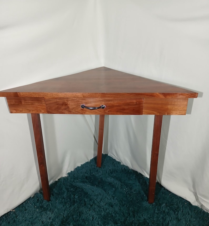 May include: A triangular wooden corner table with a drawer and a black handle. The table has a rich, reddish-brown finish and is supported by three slender legs. The table is designed to fit into a corner.