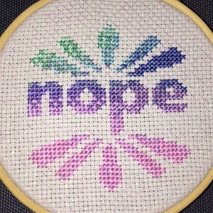 May include: A white embroidery hoop with a colorful cross-stitch design. The design features the word "hope" in a rainbow gradient and a stylized flower design.