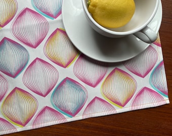 Può includere: Un tovaglietta bianca con un motivo geometrico di ovali sovrapposti in rosa, giallo e blu. La tovaglietta ha un bordo cucito giallo ed è su una superficie di legno.