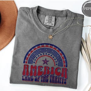 May include: A gray t-shirt with a vintage-style graphic of a rainbow with stars and the text "America Land of the Tariffs".